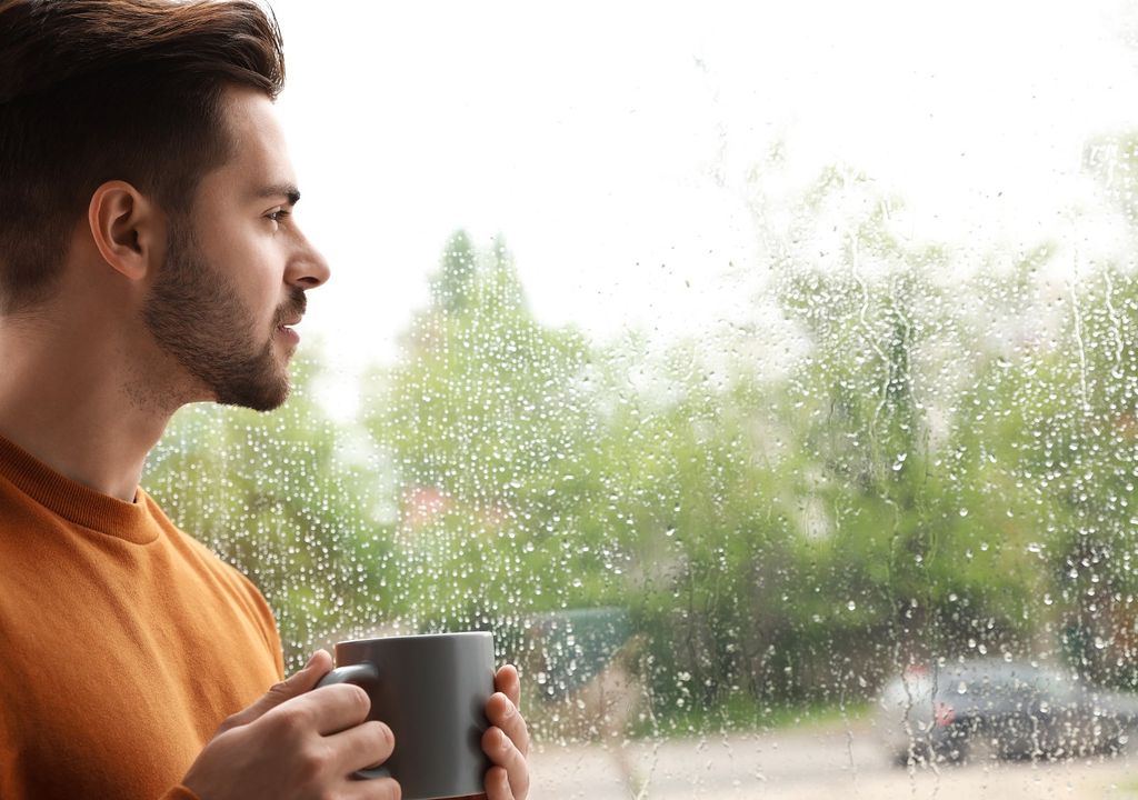 Hombre sosteniendo una taza en la mano mirando por la ventana hacia afuera en un día lluvioso