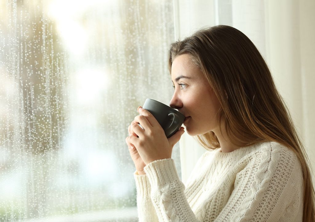 Mujer tomando bebida caliente frente a la ventana en día de lluvia