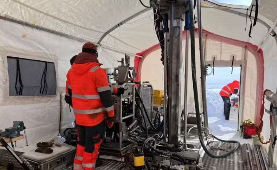 Un taladro de gran tamaño dentro de una tienda de campaña, operado por una persona vestida con un traje rojo. El taladro geológico subhielo ágil del Programa de Perforación de Hielo de la Fundación Nacional de Ciencias se utilizó para perforar más de 1600 metros de la capa de hielo de Groenlandia en la cima del domo Prudhoe para alcanzar el lecho rocoso subyacente. Foto: Jason Briner/Universidad de Buffalo