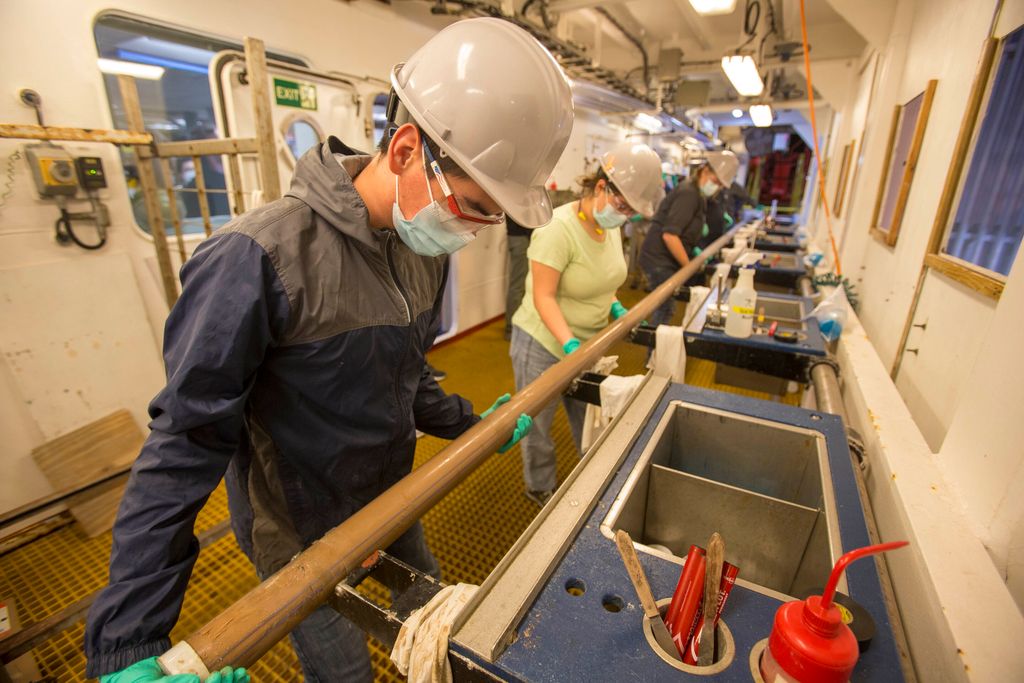 Arriba a la izquierda, Oscar Cavazos (Especialista de Laboratorio Marino, IODP JRSO) se une a otros técnicos marinos para preparar un nuevo núcleo de sedimento que se seccionará en la pasarela del JOIDES Resolution en 2022. Crédito: Erick Bravo, IODP JRSO