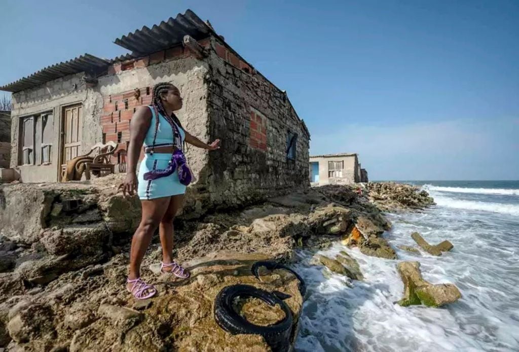 Una mujer frente a una casa antigua, mostrando cómo el aumento del nivel del mar ha erosionado la costa casi hasta los cimientos de la casa. Foto: The Conversation Una mujer frente a una casa antigua, mostrando cómo el aumento del nivel del mar ha erosionado la costa casi hasta los cimientos de la casa. Foto: The Conversation