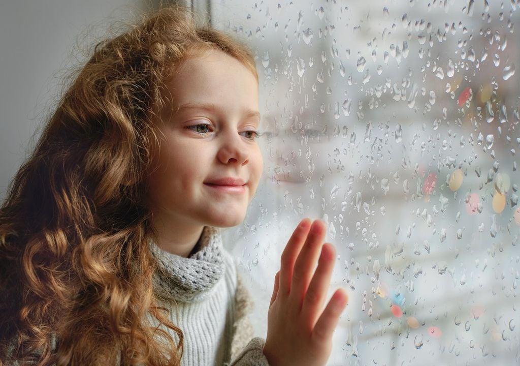 Niña mirando por la ventana hacia el exterior, hay gotas de lluvia sobre la ventana