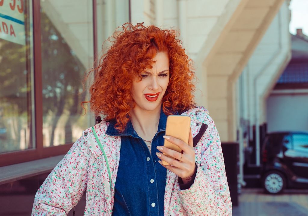 Mujer cabello rojizo con celular en mano y cara de duda