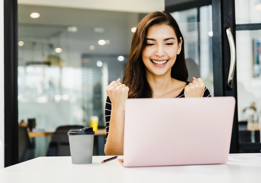 Mujer joven contenta frente al computador con café a su derecha fondo de oficina
