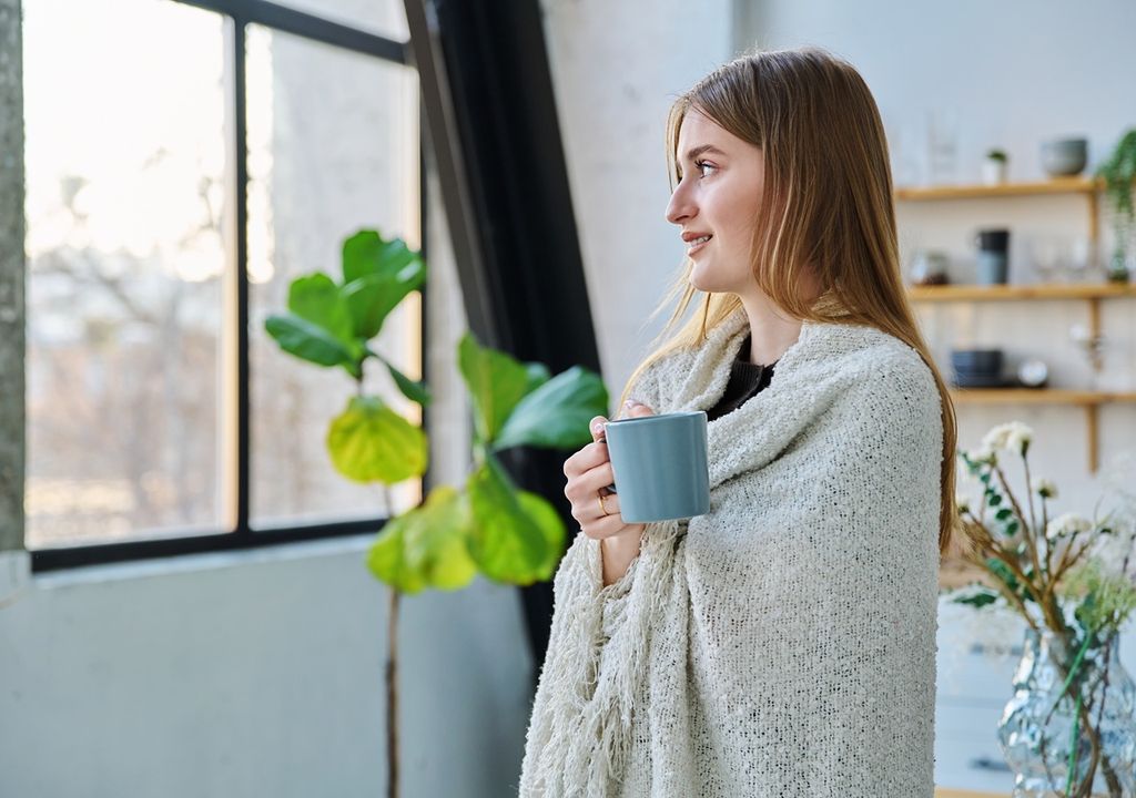 persona mirando hacia una ventana, con una taza en sus manos, y con un cobertor sobre su espalda persona mirando hacia una ventana, con una taza en sus manos, y con un cobertor sobre su espalda