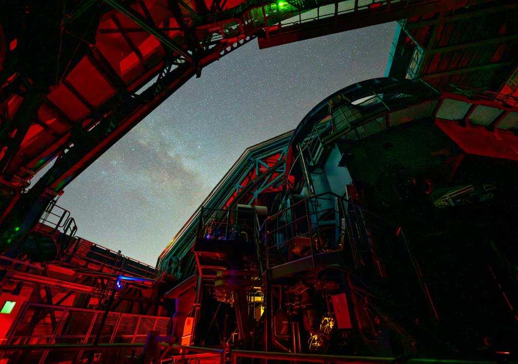 El enorme telescopio del Observatorio Vera C. Rubin se alza imponente mientras apunta hacia el cielo nocturno, a través de la abertura rectangular de su cúpula.