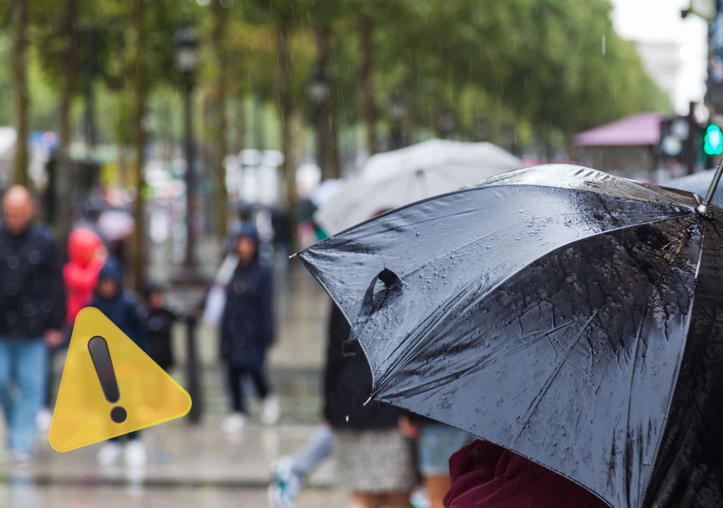 personas caminando bajo la lluvia, algunas de ellas con paraguas. Hay un símbolo de advertencia