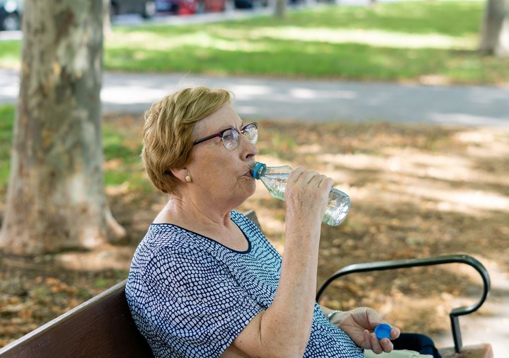 Persona adulto mayor bebiendo agua desde u na botella plástica, sentada bajo la sombra de árboles de una plaza, en un banco