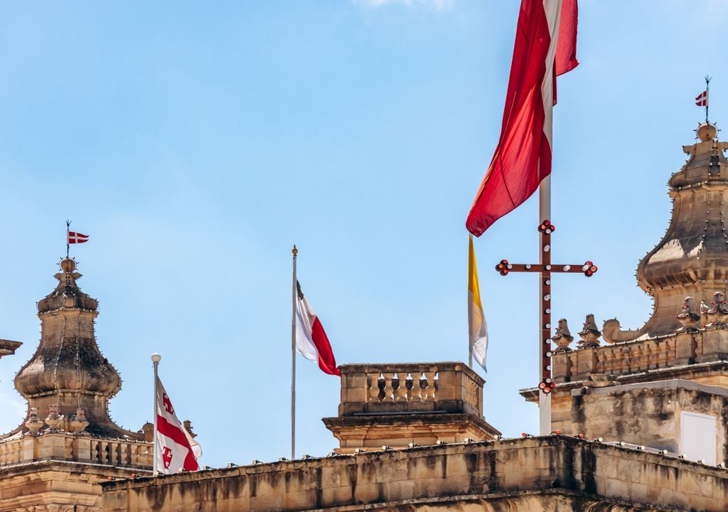 La bandera de la Soberana Orden Militar de Malta (roja con la cruz latina blanca) ondea junto a otras enseñas en un edificio histórico.