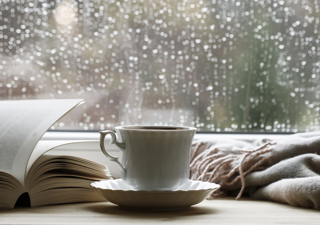 Ventana con gotas de lluvia. Una taza, un libro, y una bufanda.