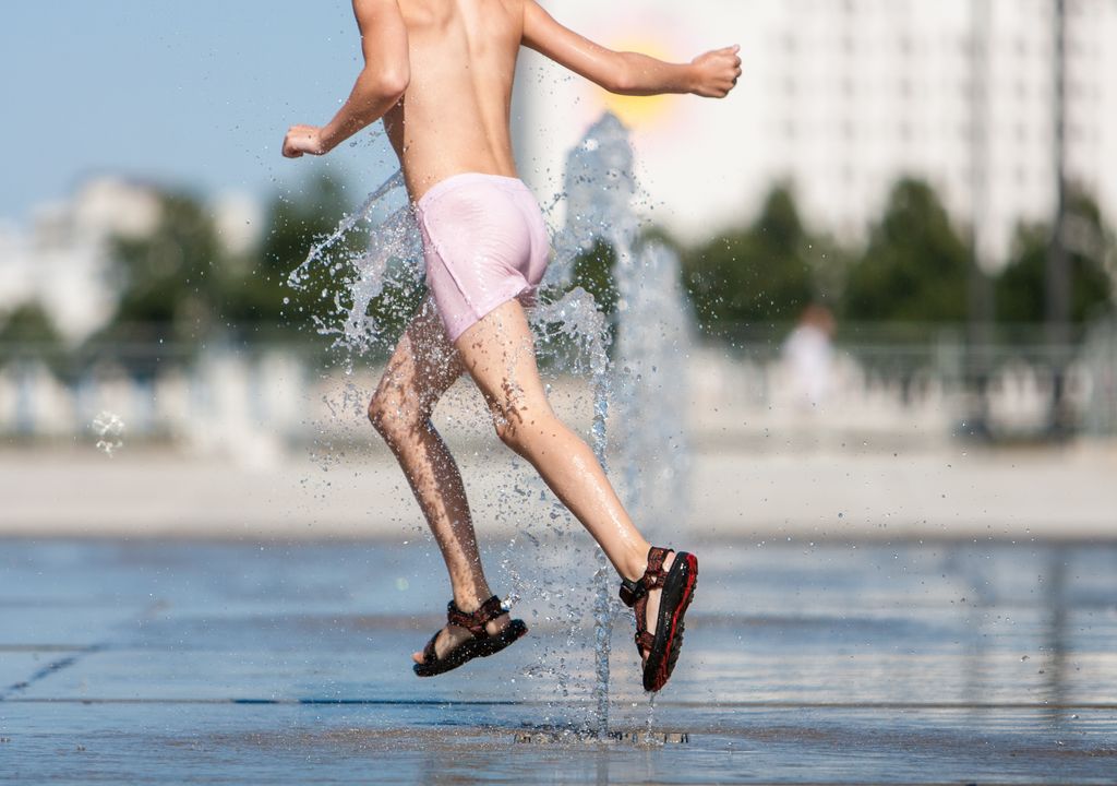 Mann mit Badehose und Sandalen springt durch eine Wasserfontäne. Schwülheiß sollen die Temperaturen Ende Juli in Nordrhein-Westfalen werden.