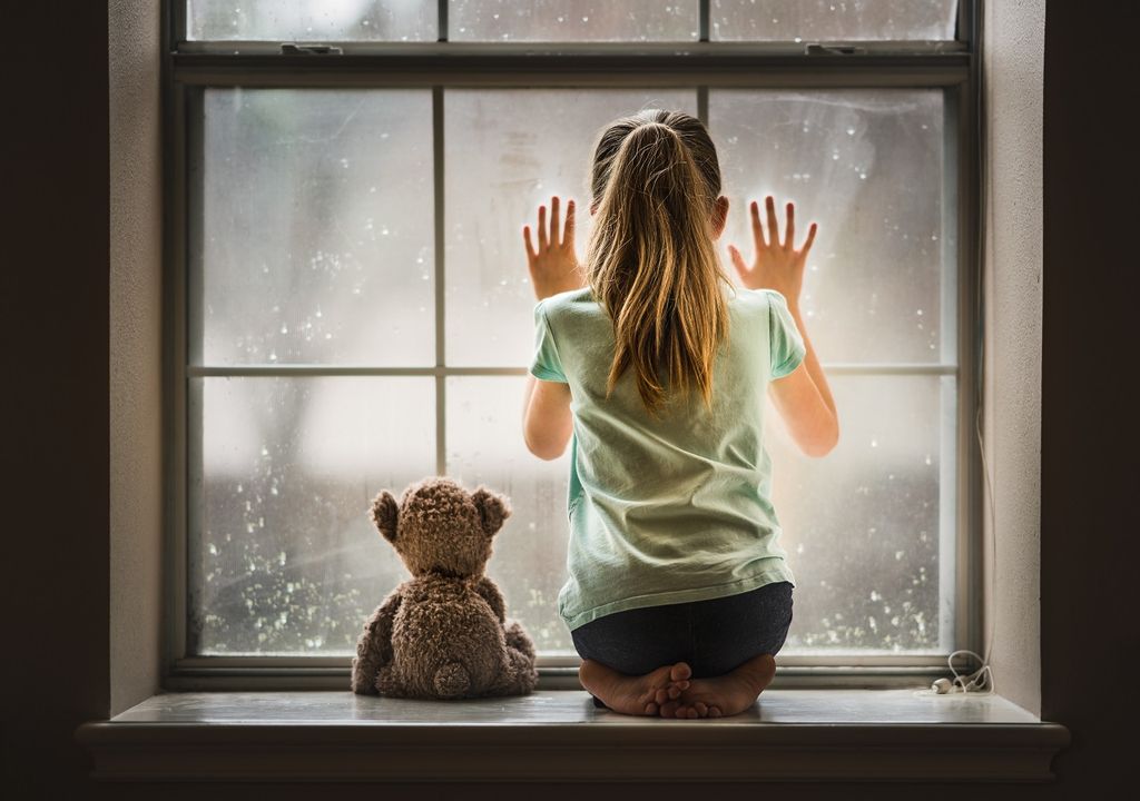 Niña con su oso mirando la lluvia desde ventana
