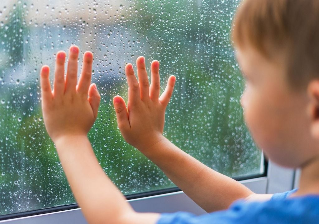 Niño con manos en la ventana, mirando la lluvia afuera