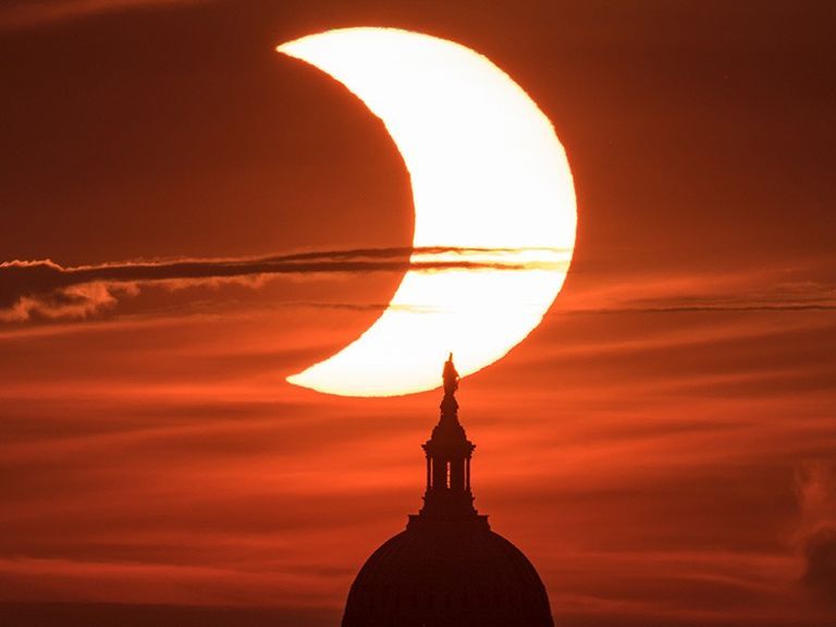 Se muestra un cielo rojizo con el brillante círculo del Sol, del que se ha extraído una parte oscura. El círculo oscuro que cubre aproximadamente un tercio del lado izquierdo del Sol es la Luna, durante un eclipse solar. En primer plano se divisa la silueta del Capitolio de los Estados Unidos.El 10 de junio de 2021, un eclipse solar parcial emerge desde detrás del Capitolio de los Estados Unidos, visto desde Arlington, Virginia. NASA/Bill Ingalls