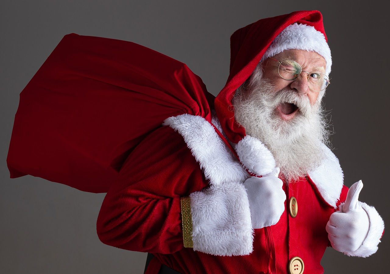 Papai Noel segurando seu saco de presentes sobre o ombro, olha para a camera e dá uma piscadela