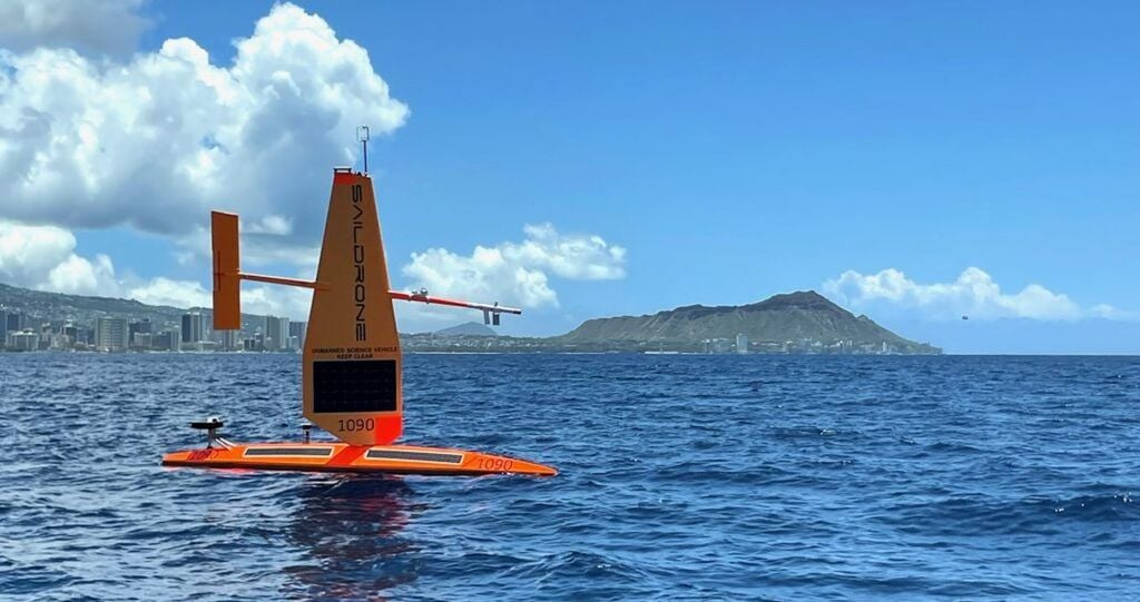 Un dron Saildrone naranja atraviesa una bahía con una montaña en la distancia. El Saildrone Explorer SD-1090 zarpa de Honolulu el 21 de agosto de 2026 para una misión de cuatro meses al Pacífico ecuatorial, a 2400 kilómetros al sur. Cuatro Saildrones capturarán datos oceánicos y atmosféricos para los científicos del Laboratorio Ambiental Marino del Pacífico de la NOAA. Foto cortesía de Saildrone.