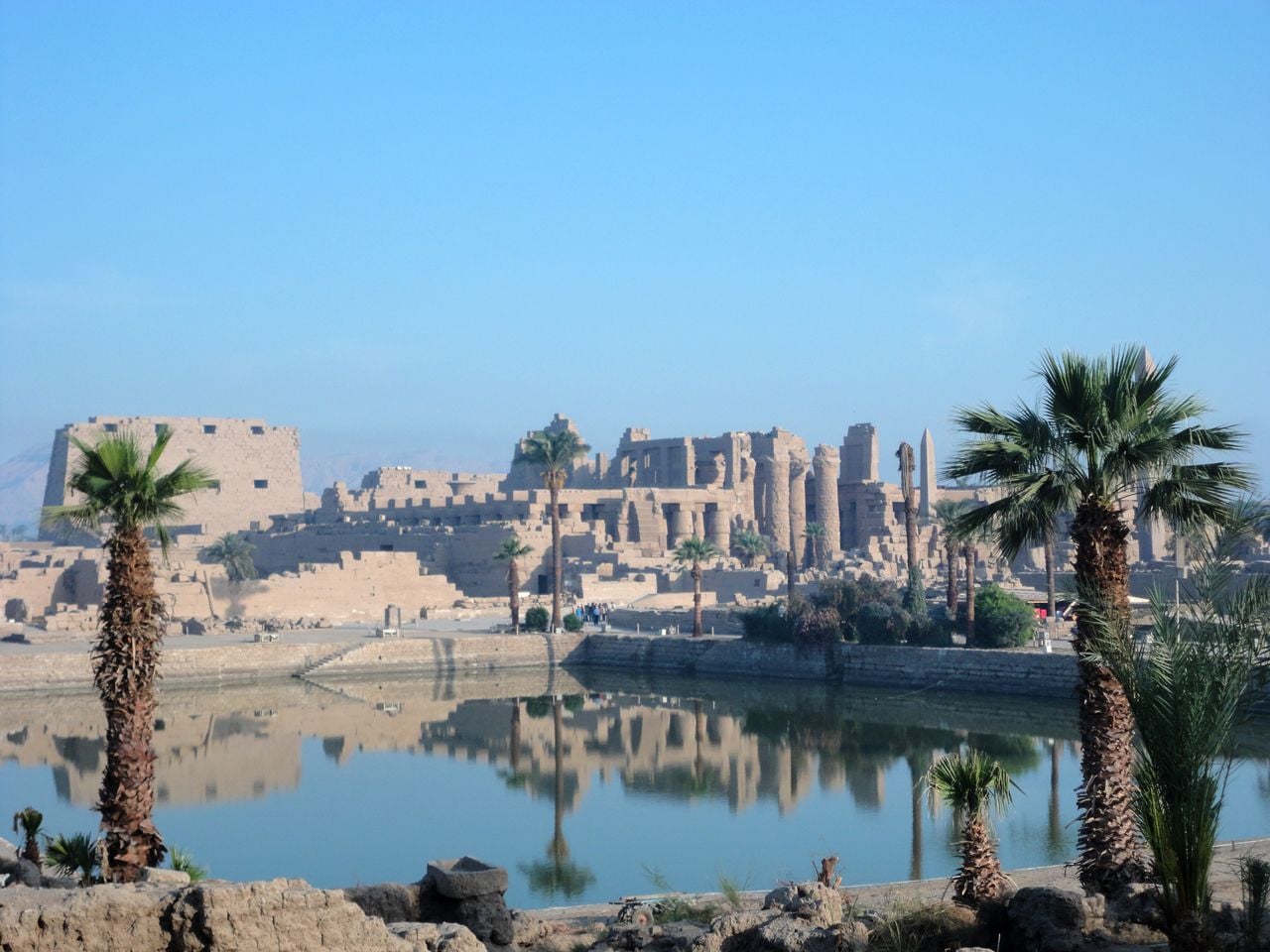 Ruinas antiguas del complejo del templo de Karnak en Luxor, Egipto, vistas a través de un gran estanque con palmeras en primer plano. Las columnas de piedra, los pilonos y los obeliscos se reflejan en las tranquilas aguas bajo un cielo azul despejado. Crédito: Dr. Ben Pennington