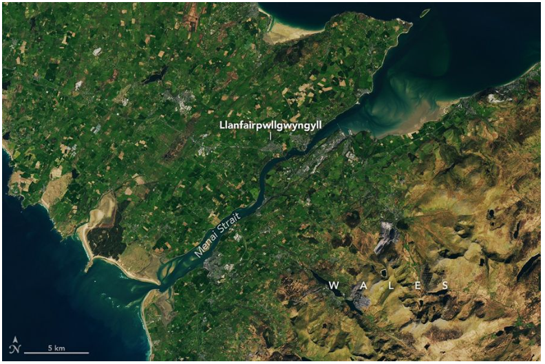 Una vista más amplia de la misma zona muestra cómo el estrecho de Menai se conecta con el océano abierto al norte y al sur de la isla de Anglesey. El lado derecho de la imagen presenta un terreno más montañoso con menos tierras de cultivo. NASA