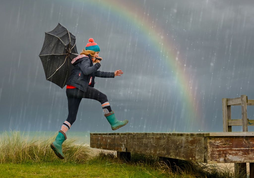 Mujer con paraguas saltando hacia plataforma de madera fondo con arcoiris y nube oscura