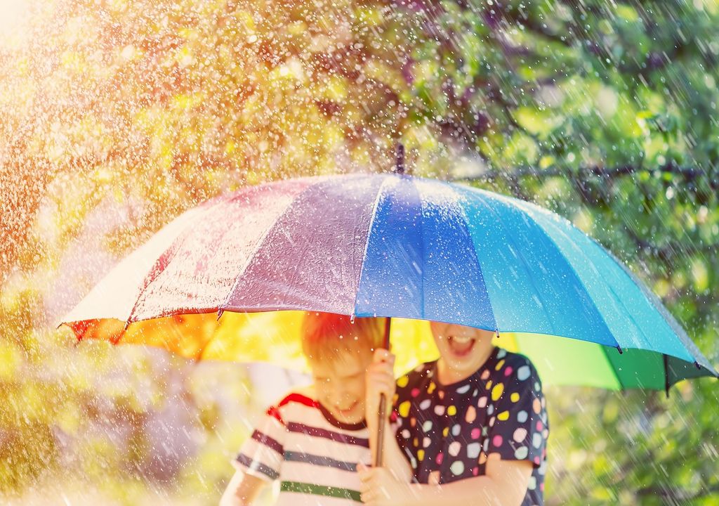 Niño y niña con paraguas multicolor bajo la lluvia fondo soleado con árboles