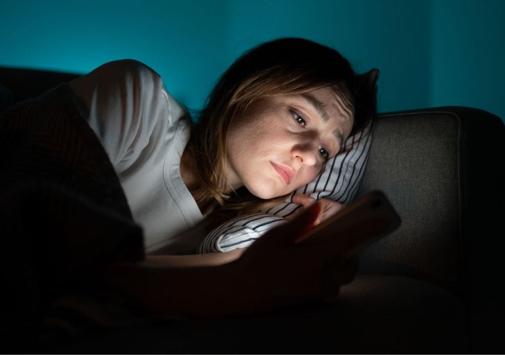 Joven en un sofá a oscuras con cara de ansiedad, celular ilumina su rostro.