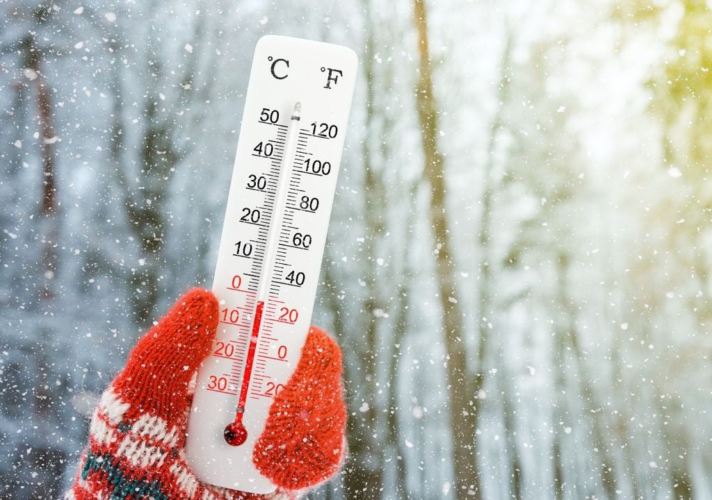 Mano con guante de lana rojo sujetando termómetro fondo con árboles y nieve