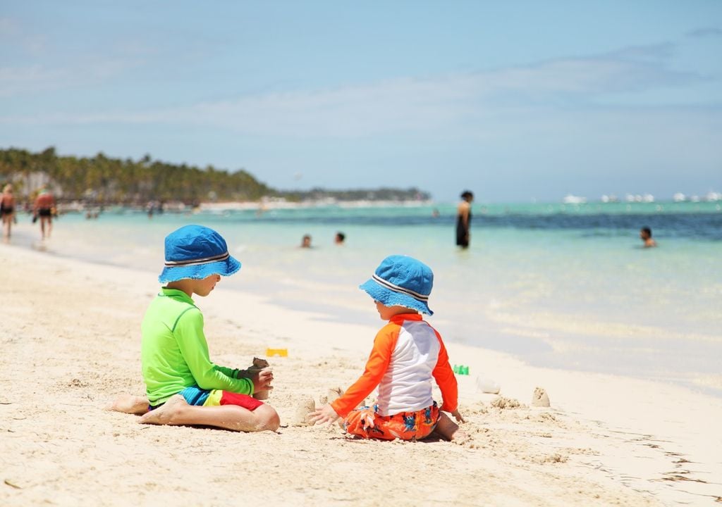 Crianças na praia, brincando na areia, vestindo camisas de manga comprida com proteção UV e chapéus