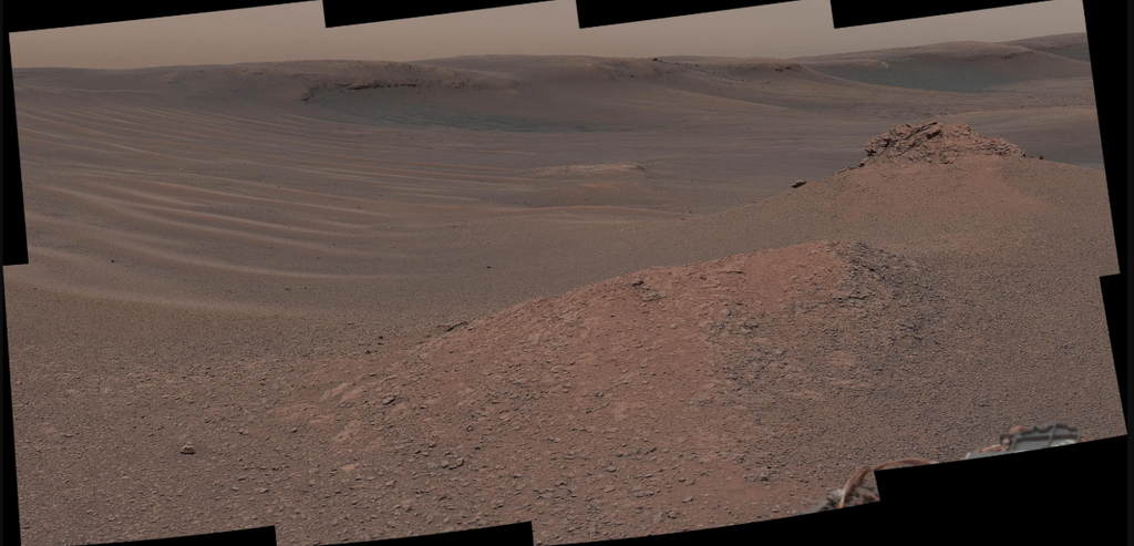 Un vasto y desolado paisaje marciano presenta ondulaciones de arena barridas por el viento bajo un cielo brumoso de color salmón, con formaciones rocosas irregulares en primer plano y colinas onduladas de color marrón rojizo al fondo. La cámara Mastcam del rover Curiosity capturó este mosaico el 3 de febrero de 2019, de una región del monte Sharp con abundantes rocas arcillosas que se formaron cuando había lagos y arroyos hace miles de millones de años. La muestra "Mary Anning 3" se encontró en esta región rica en arcilla. NASA/JPL-Caltech/MSSS