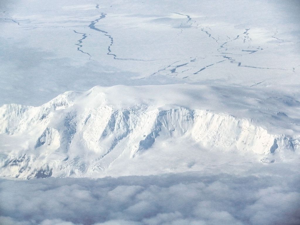 En una fotografía tomada desde un avión con vista oblicua, se aprecia una isla escarpada cubierta de hielo, parcialmente velada por las nubes y rodeada de hielo marino fracturado. La foto fue tomada desde el laboratorio científico aerotransportado DC-8 de la NASA durante un vuelo de la Operación IceBridge el 3 de noviembre de 2011, la isla Pedro I se encuentra enclavada entre el hielo marino y las nubes.Fotografía cortesía de Christopher Shuman, UMBC (jubilado).