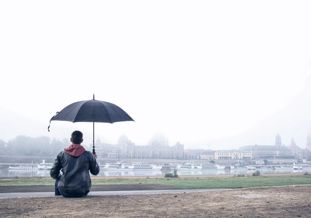 Ein Mann sitzt mit Regenschirm am Elbufer und blickt Richtung Dresdener Altdtadt. Es ist trüb und regnerisch.