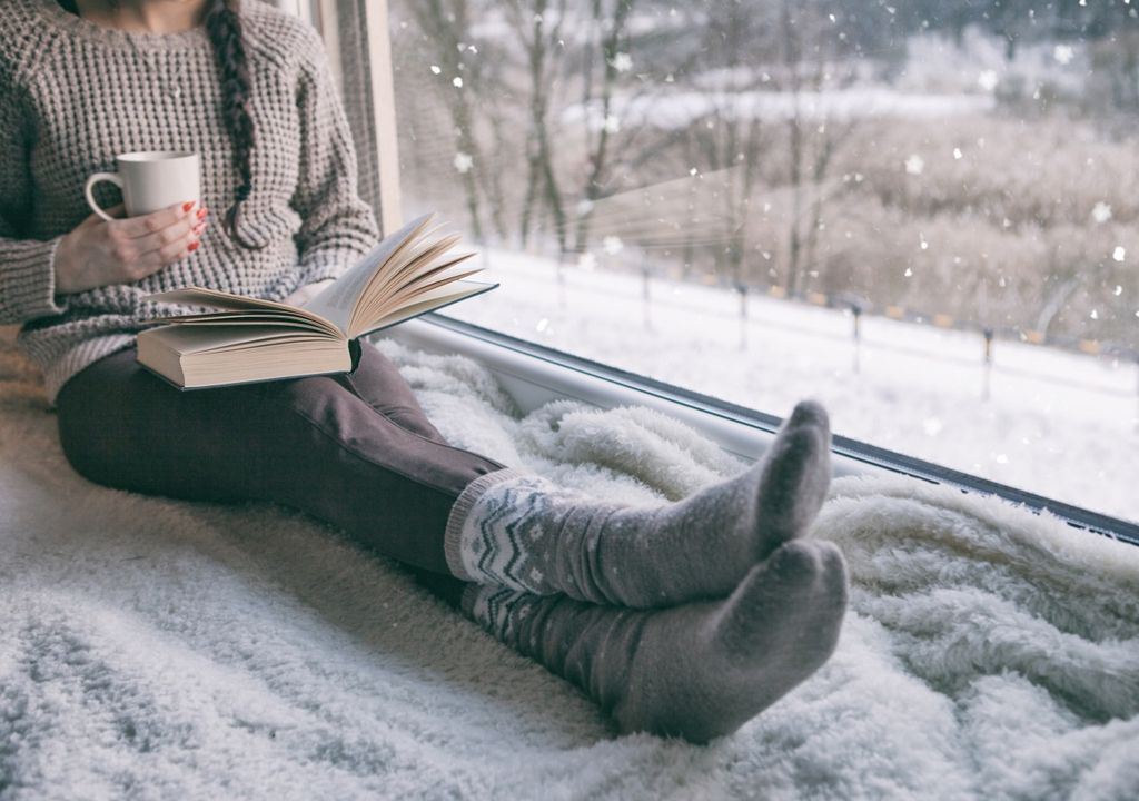 Mujer sentada cerca de la ventana leyendo un libro y una taza en la mano; paisaje de nieve en el exterior